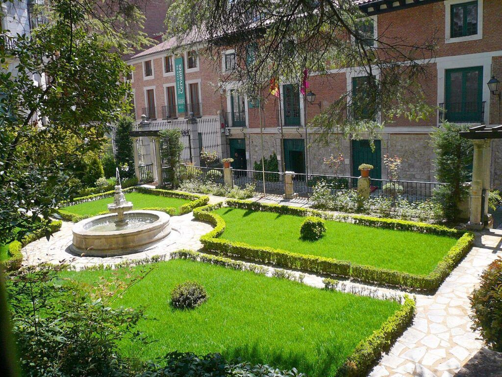 Jardín de entrada del Museo Casa de Cervantes en Valladolid, con una fuente, parterres de césped y la fachada de ladrillo al fondo