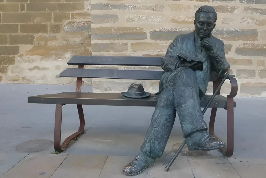 Escultura de bronce de Antonio Machado sentado en un banco leyendo un libro, con su sombrero y su bastón, en Baeza