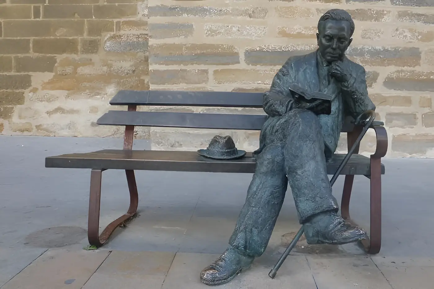 Escultura de bronce de Antonio Machado sentado en un banco leyendo un libro, con su sombrero y su bastón, en Baeza